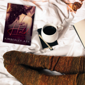 On a rumpled white cover, a person with thick brown wool socks sits will a cup of coffee, a notebook, and a paperback copy of Hold.